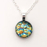 A round pendant necklace featuring an iridescent, mosaic-like pattern with gold, blue, green, and purple hues, suspended from a silver link chain on a plain white background.